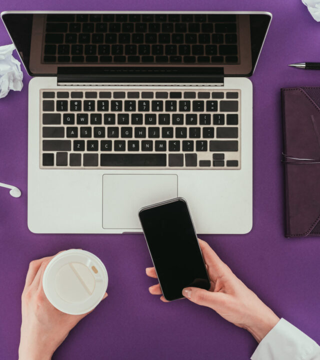 cropped shot of businesswoman using smartphone and drinking coffee at workplace on purple surface